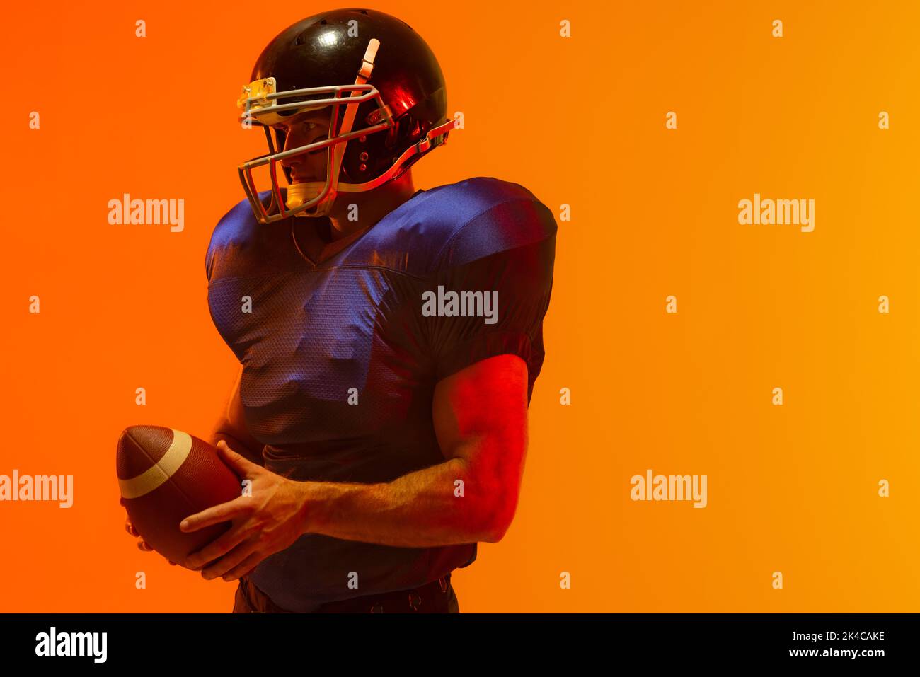 Caucasian male american football player holding ball with neon orange lighting. Sport, movement, training and active lifestyle concept. Stock Photo