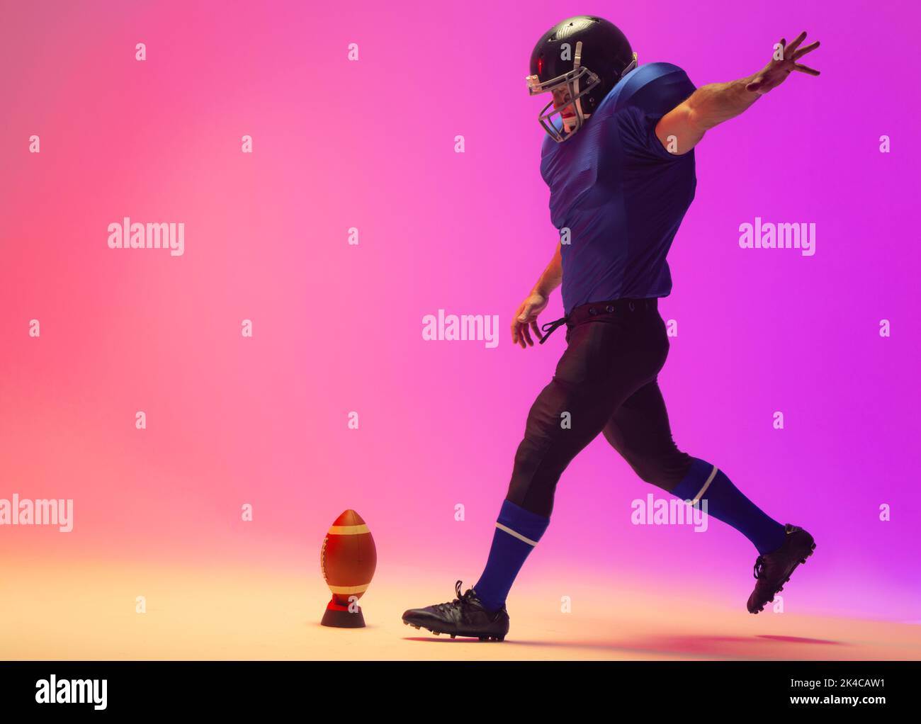 Caucasian male american football player kicking ball with neon pink lighting. Sport, movement, training and active lifestyle concept. Stock Photo