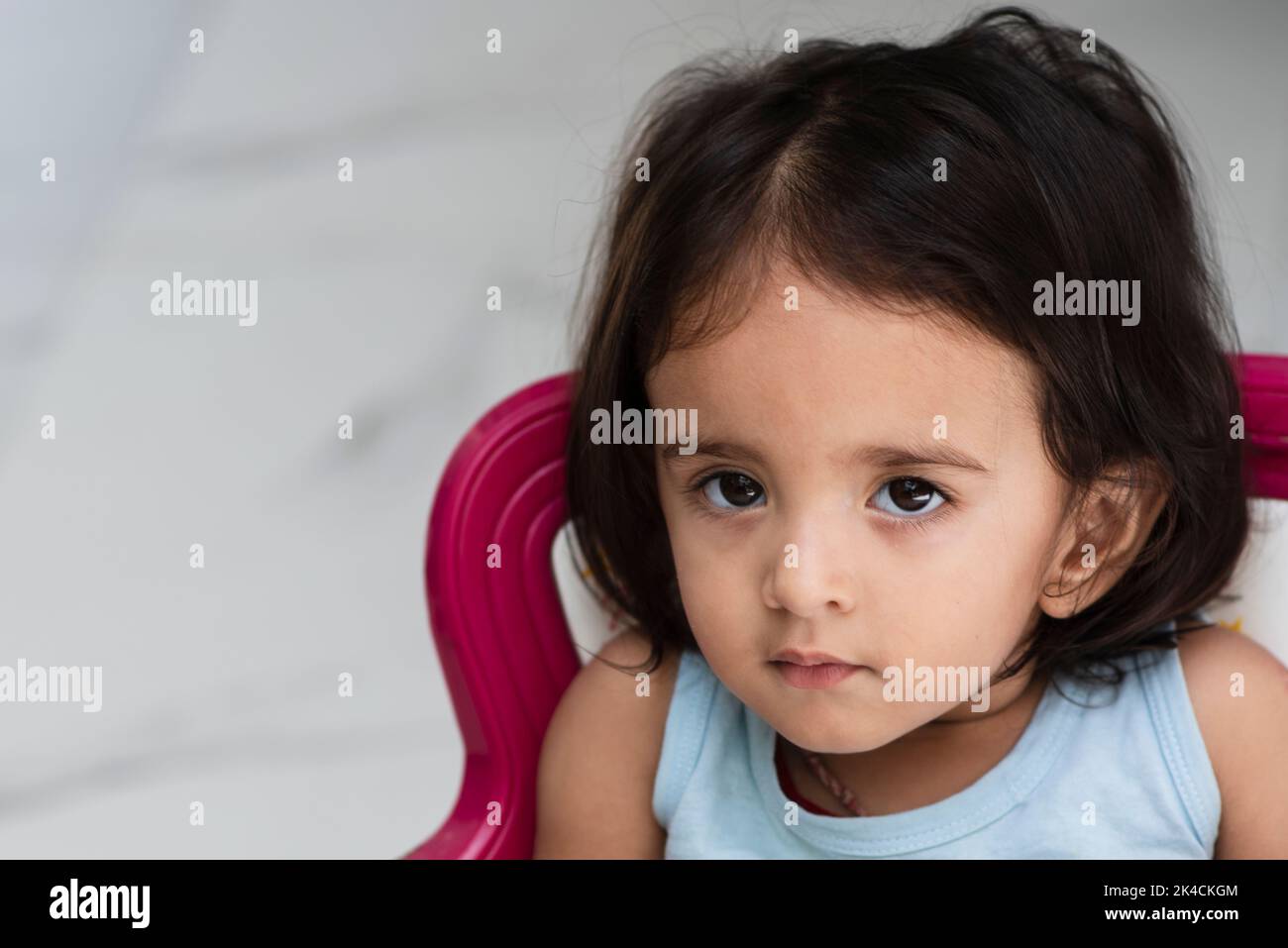 Cute and adorable Indian baby girl wearing aqua color t shirt sitting on a baby pink color chair looking up and giving innocent expressions Stock Photo Alamy