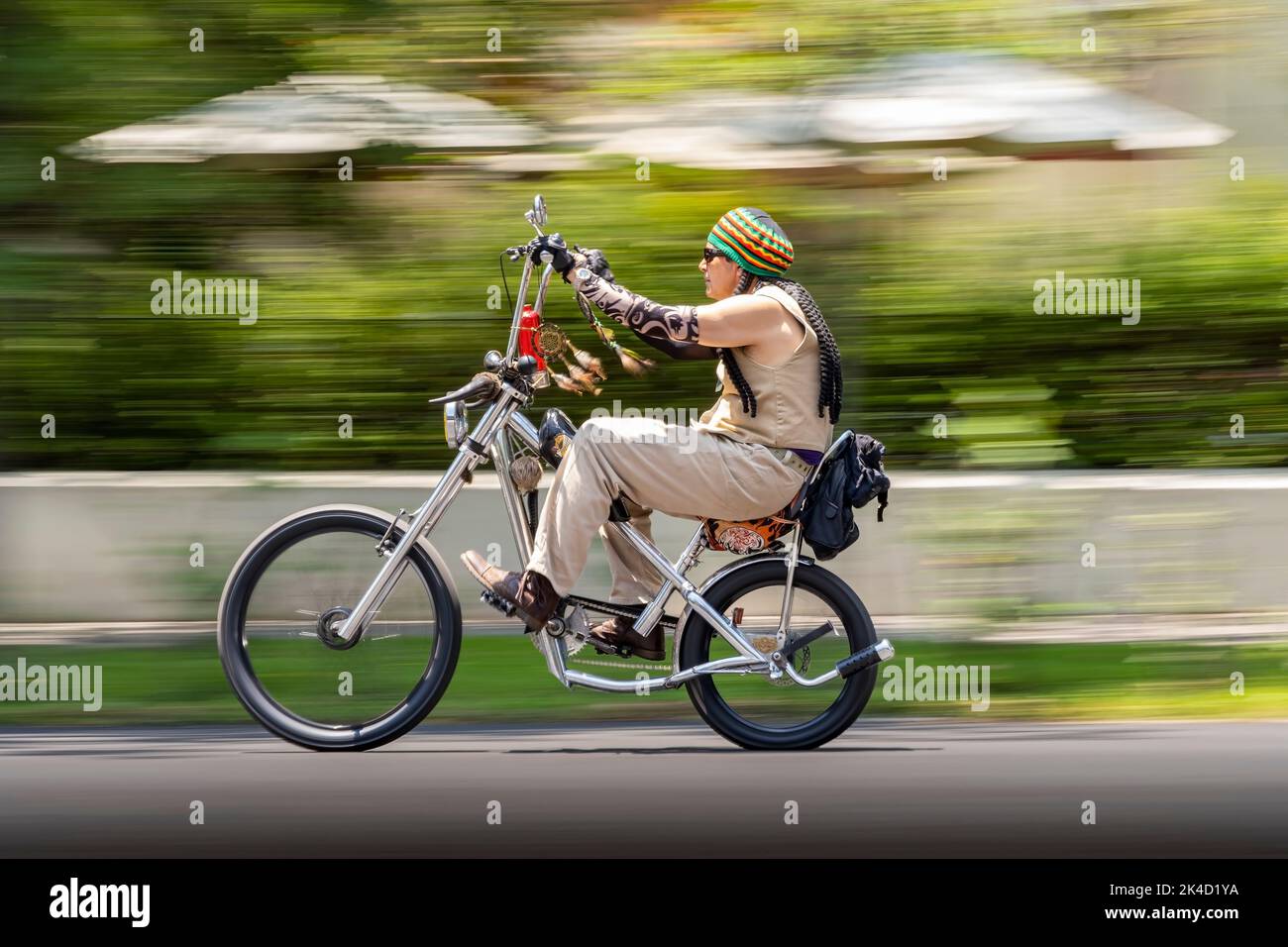 man bicycle, with dreadlocks and Rastafarian cap, sleeve simulating tattoos, custom chopper bike, blurred motion Stock Photo