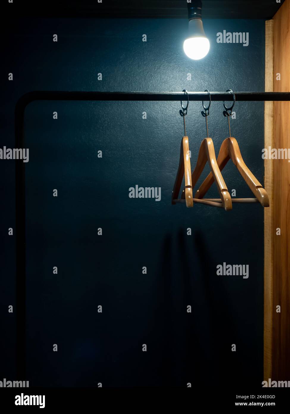 Three empty wooden clothes hangers without shirts or dress hanging on a black cloth rack with light bulb in the wood wardrobe on dark blue background Stock Photo