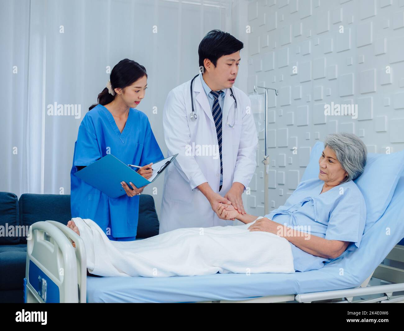 Asian male doctor, professional in white lab coat holding elderly patient's hand for check to examine heartbeat pulse and nursing assistant taking not Stock Photo