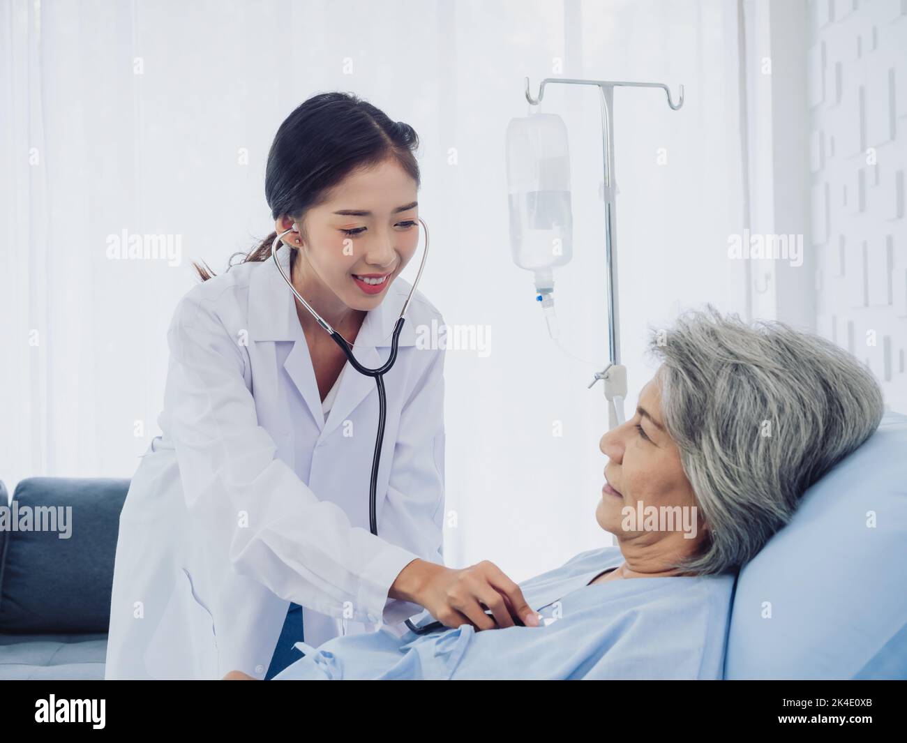 A beautiful young Asian woman doctor in white suit smiled while using stethoscope to examine, listen to heartbeat of elderly old female patient in blu Stock Photo