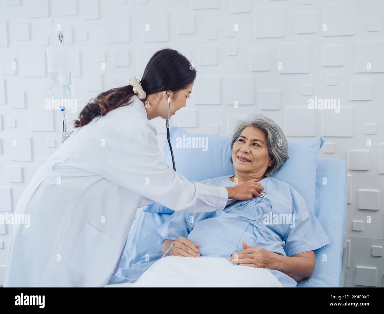 Happy smile Asian elderly old female patient in light blue dress lying on bed while beautiful young woman doctor in white suit using stethoscope to ex Stock Photo