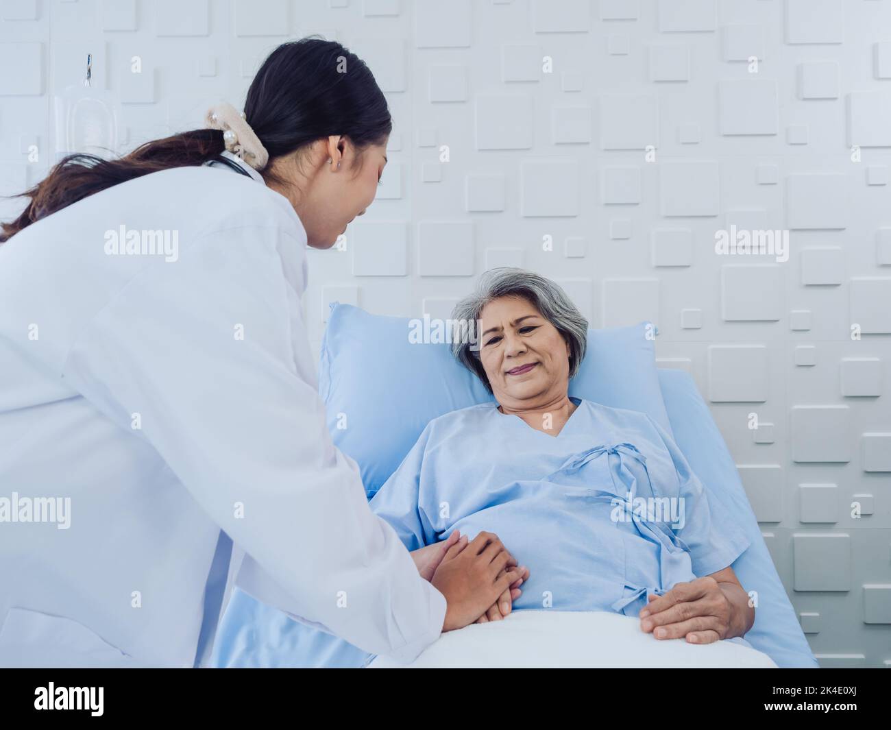 Happy Asian elderly senior woman patient in light blue dress lying on bed in saline solution was encouraging by young female doctor in white suit with Stock Photo