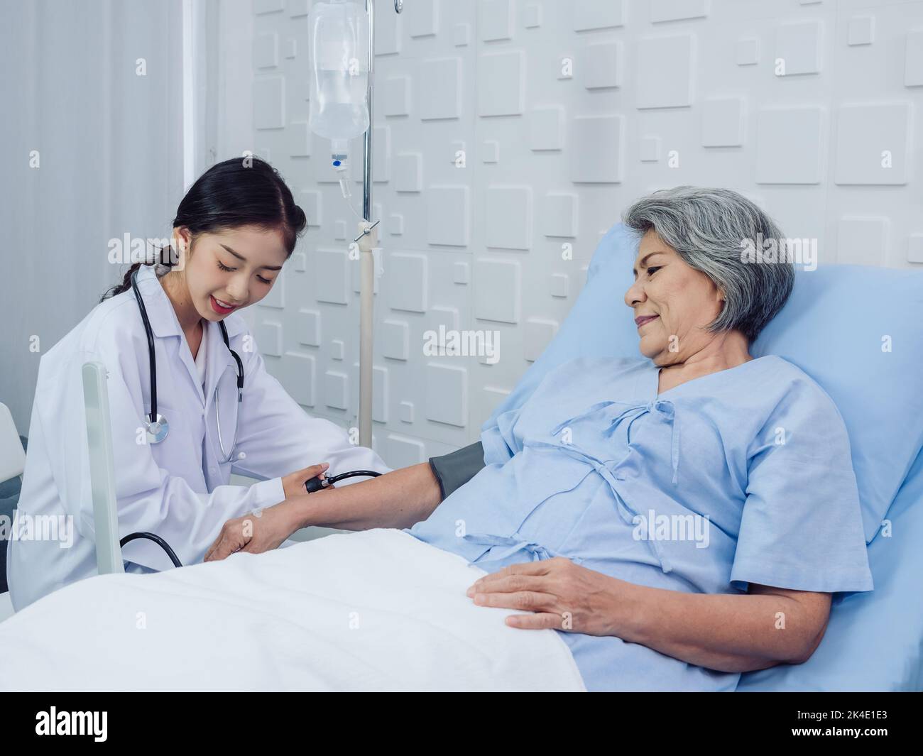 Happy smile Asian elderly old female patient in light blue dress lying on bed while beautiful young woman doctor in white suit measuring blood pressur Stock Photo