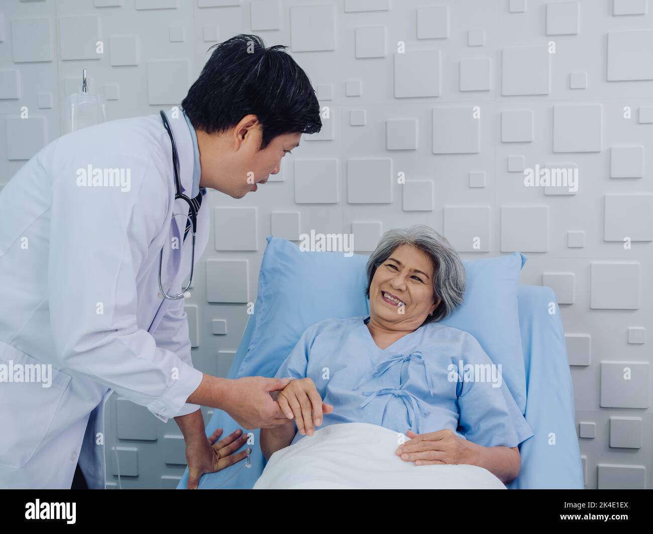 The kind Asian male doctor in white suit visits, talks and gives support, holding hand of happy elderly senior woman patient in light blue dress lying Stock Photo