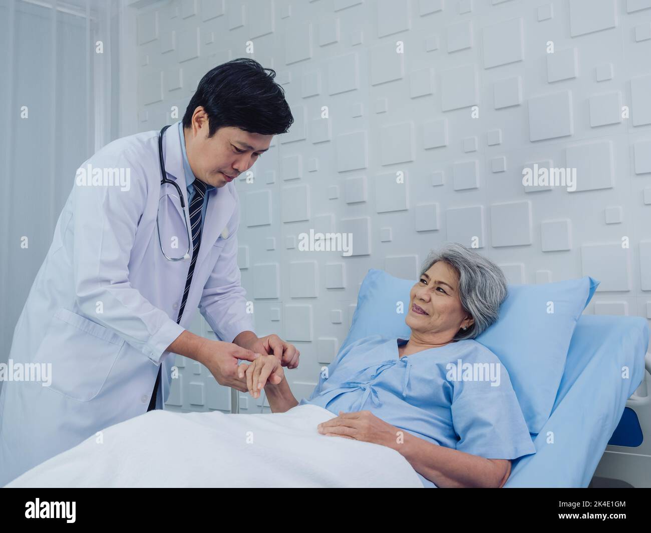 Happy smile beautiful Asian elderly old woman patient in light blue dress lying on bed while male doctor in white suit holding her hand and giving int Stock Photo