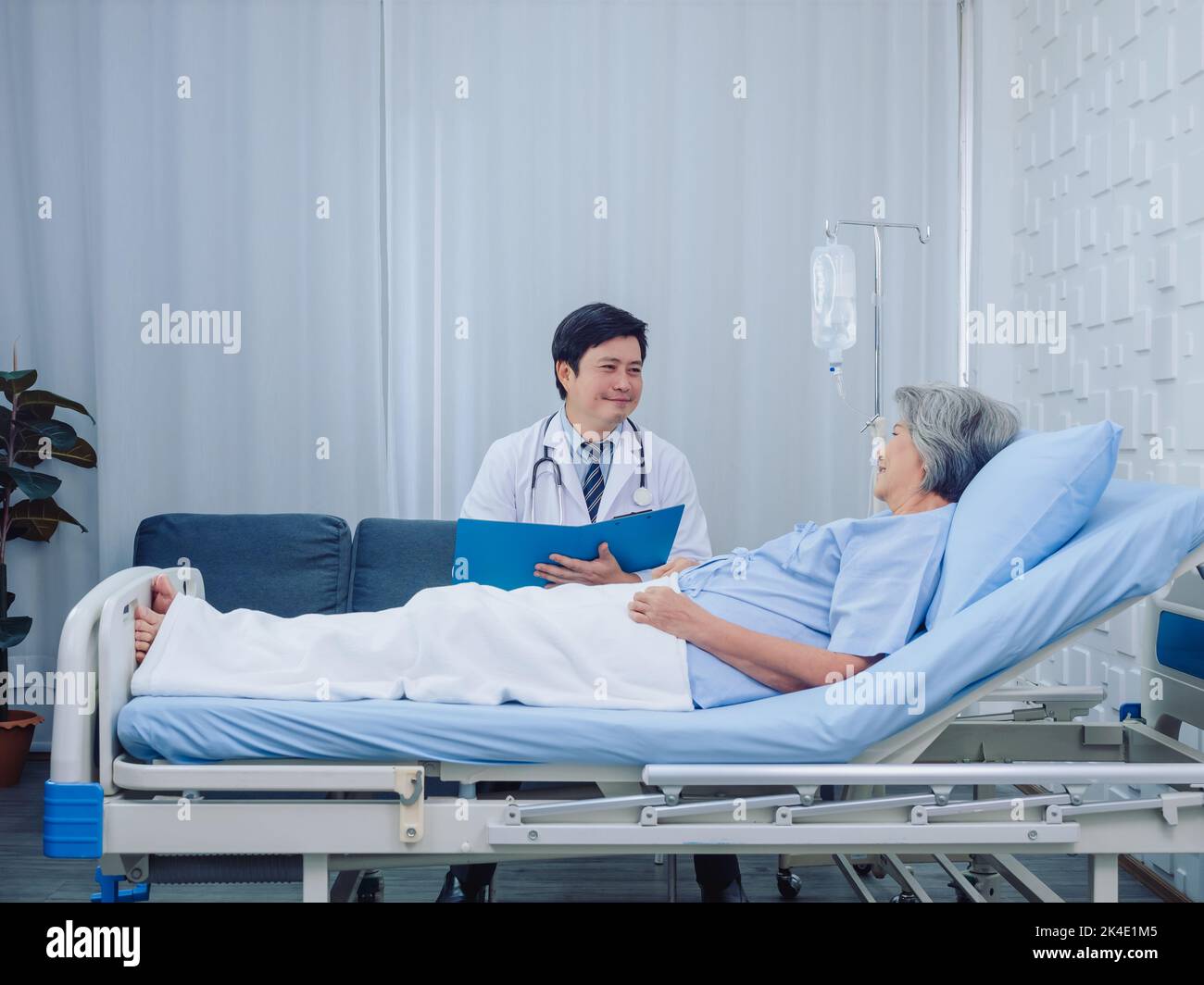 Smiling Asian doctor in white suit take notes while discussing happy Asian elderly, senior woman patient who lying on bed with receiving saline soluti Stock Photo