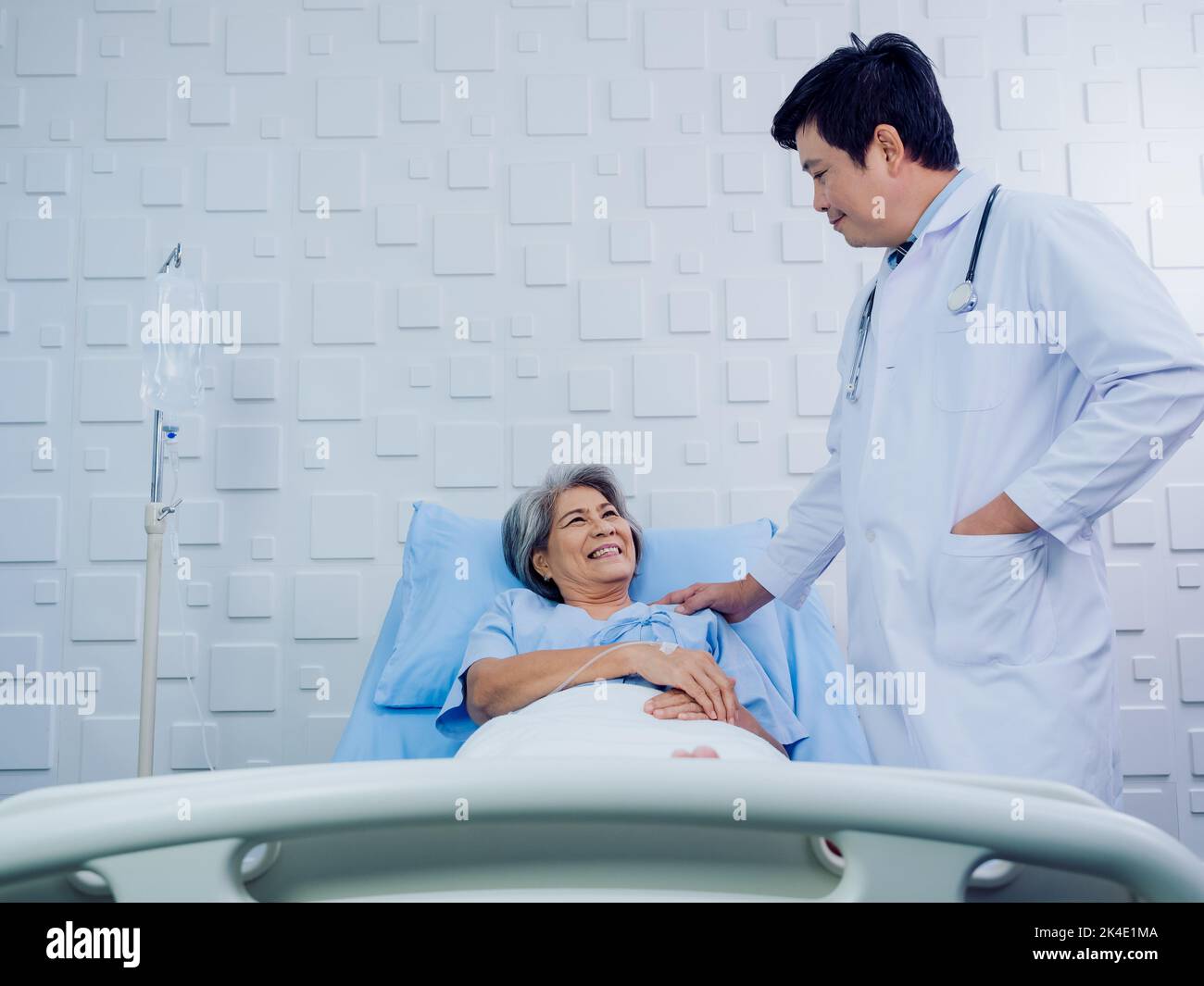 Happy smile Asian elderly, senior woman patient in light blue dress lying on bed with receiving saline solution while doctor in white touching her sho Stock Photo