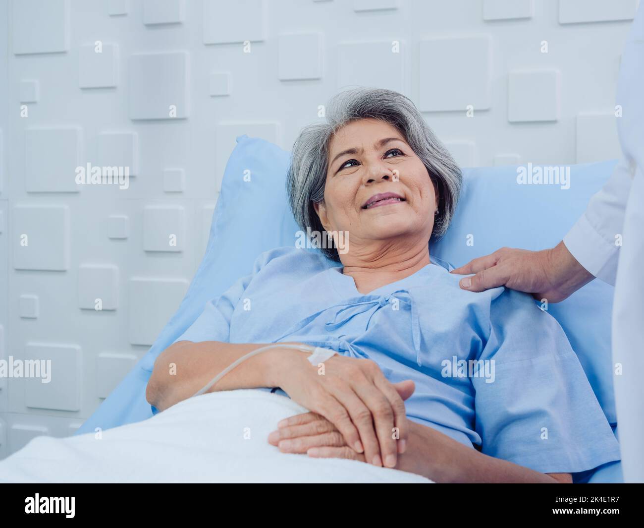 Closeup portrait of smiling Asian elderly, senior woman patient in light blue dress lying on bed while male doctor in white suit touching her shoulder Stock Photo