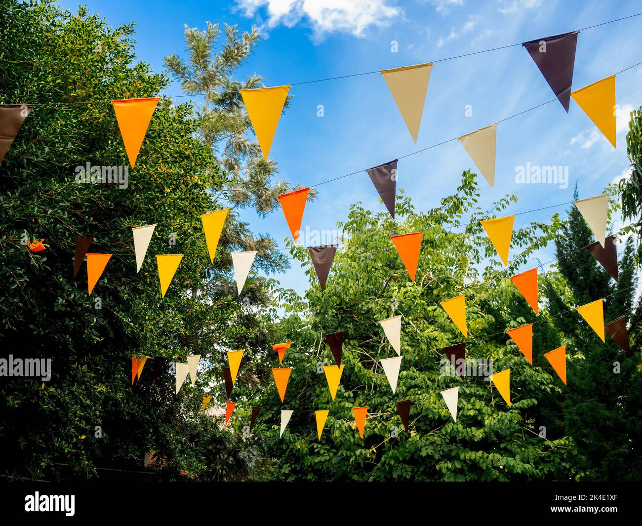 Event outdoor party ornamented with orange, yellow, brown, and white color tone flags in green garden on blue sky background. Colorful triangular flag Stock Photo