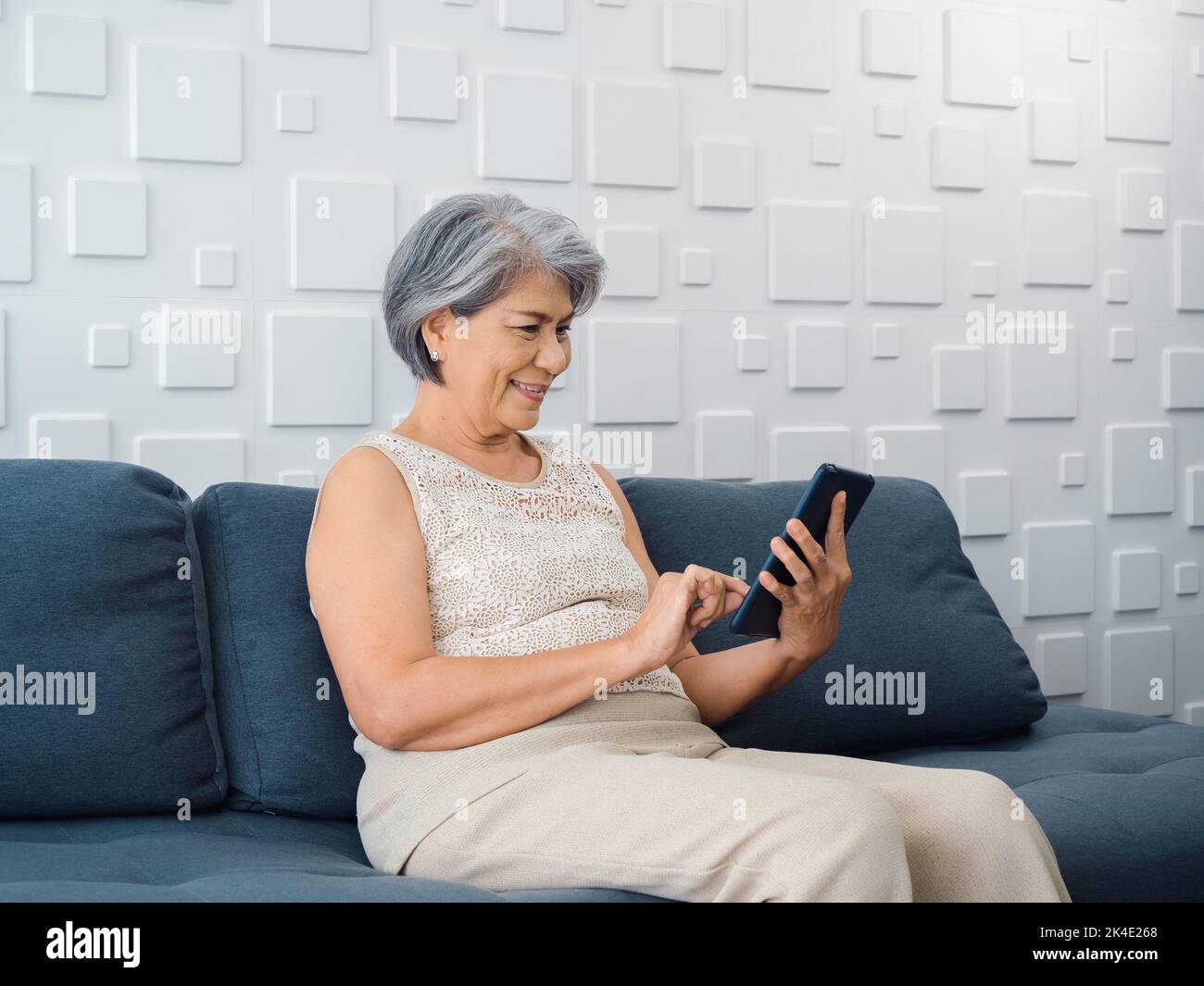 Happy Asian senior woman sit on couch, holding and looking at mobile phone screen in hand in living room. Elderly female using smart device, computer Stock Photo