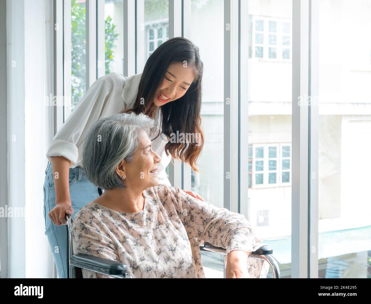 Happy Asian elderly woman, mother or grandparents on wheelchair taking care by caregiver, smiling young female, daughter or grandchild supporting at h Stock Photo
