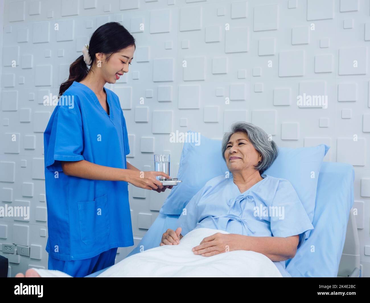 Friendly smiling young Asian nurse in blue scrub holding tray of pills for elderly patient lying on the bed in hospital room, taking medicine or vitam Stock Photo