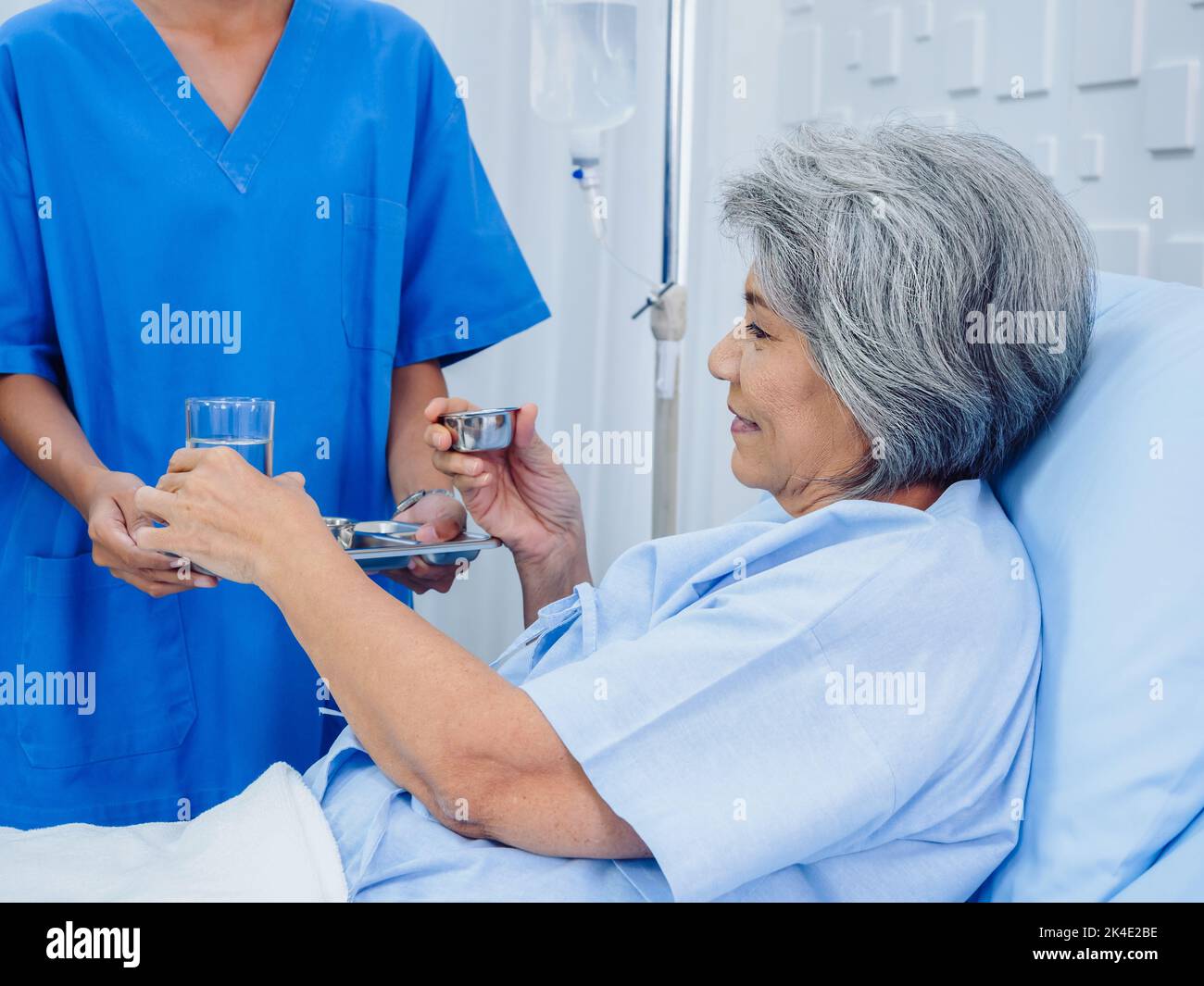 Asian elderly patient taking medicine, hold drinking water glass and pill from young nurse in blue scrub suit in hospital room, daily medicine or vita Stock Photo