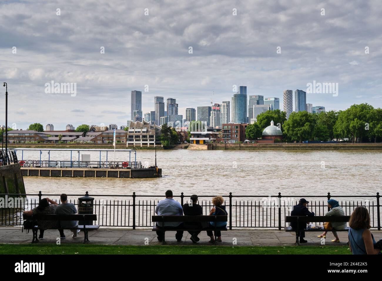 On a warm late spring day, some people sit on the benches along the Thames side in Greenwich. In background Canary Wharf skyline landscape. Stock Photo
