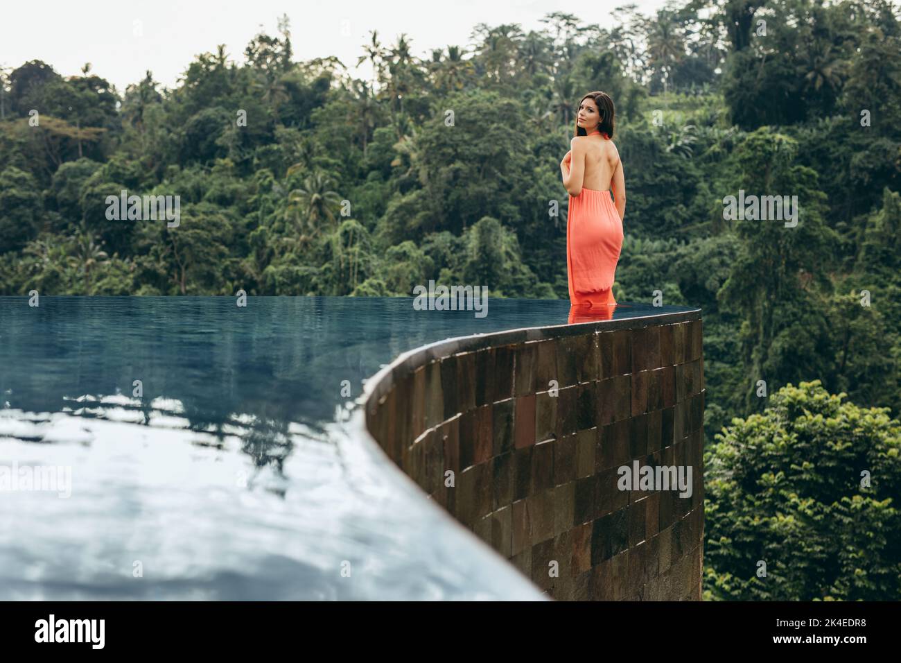 Shot of attractive young woman standing in infinity pool of holiday resort. Caucasian female model in orange dress looking over shoulder. Stock Photo