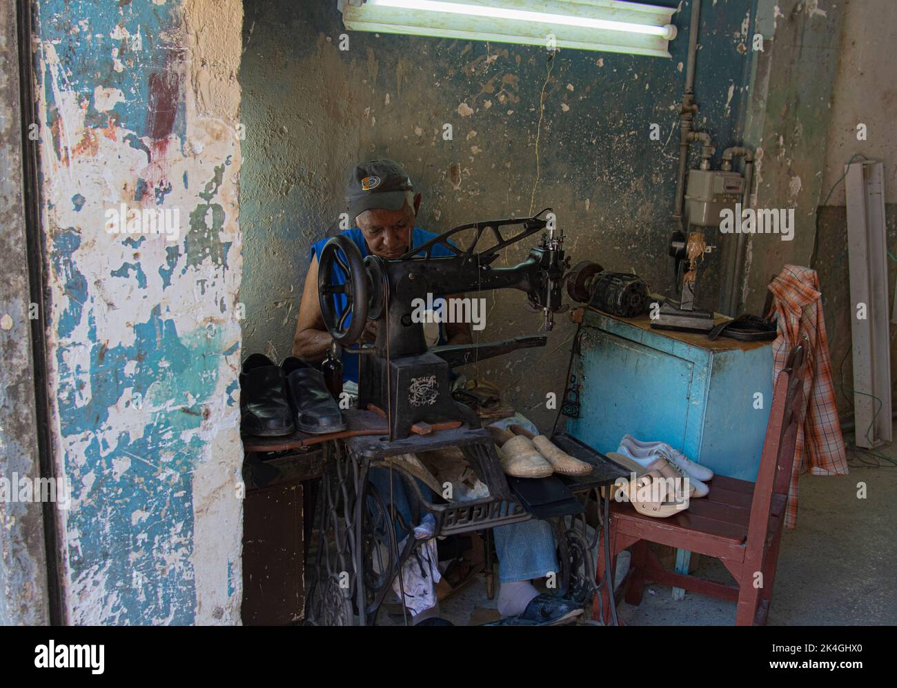 Middle aged man wearing a cap sits working on an old antiquated Singer sewing machine on mending shoes while working in a factory in Havana, Cuba. Stock Photo