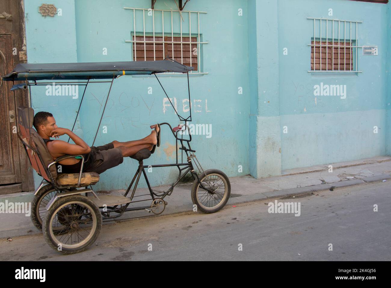 Young man resting on top of his bicycle in front of a house with a tribute to the late Cuban President Fidel Castro written on the wall. Stock Photo