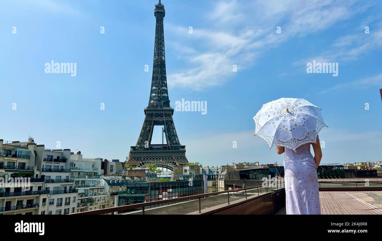 a beautiful young girl in a summer dress stands against the backdrop of the Eiffel Tower with a lace umbrella, she raised her hand and as if taking off there is a place for advertising travel agency. Stock Photo