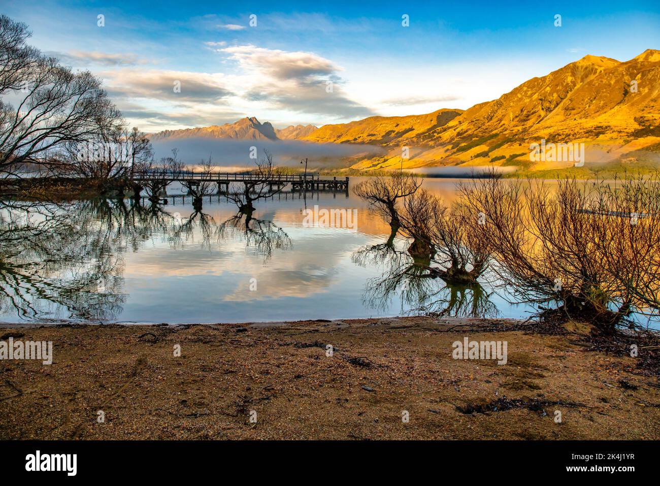 Glenorchy at sunrise as the early morning golden sun warms the mountains and the mountains and willow trees are reflected on Lake Wakatipu Stock Photo