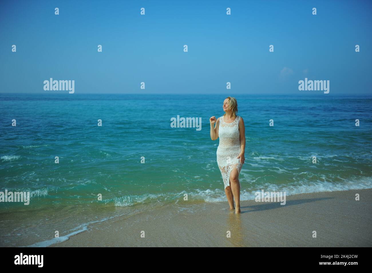 A woman of 30-40 years old in a white lace dress stands on the ocean, and waves of clear water reach her feet with a sea of different colors from turquoise to dark. High quality photo Stock Photo