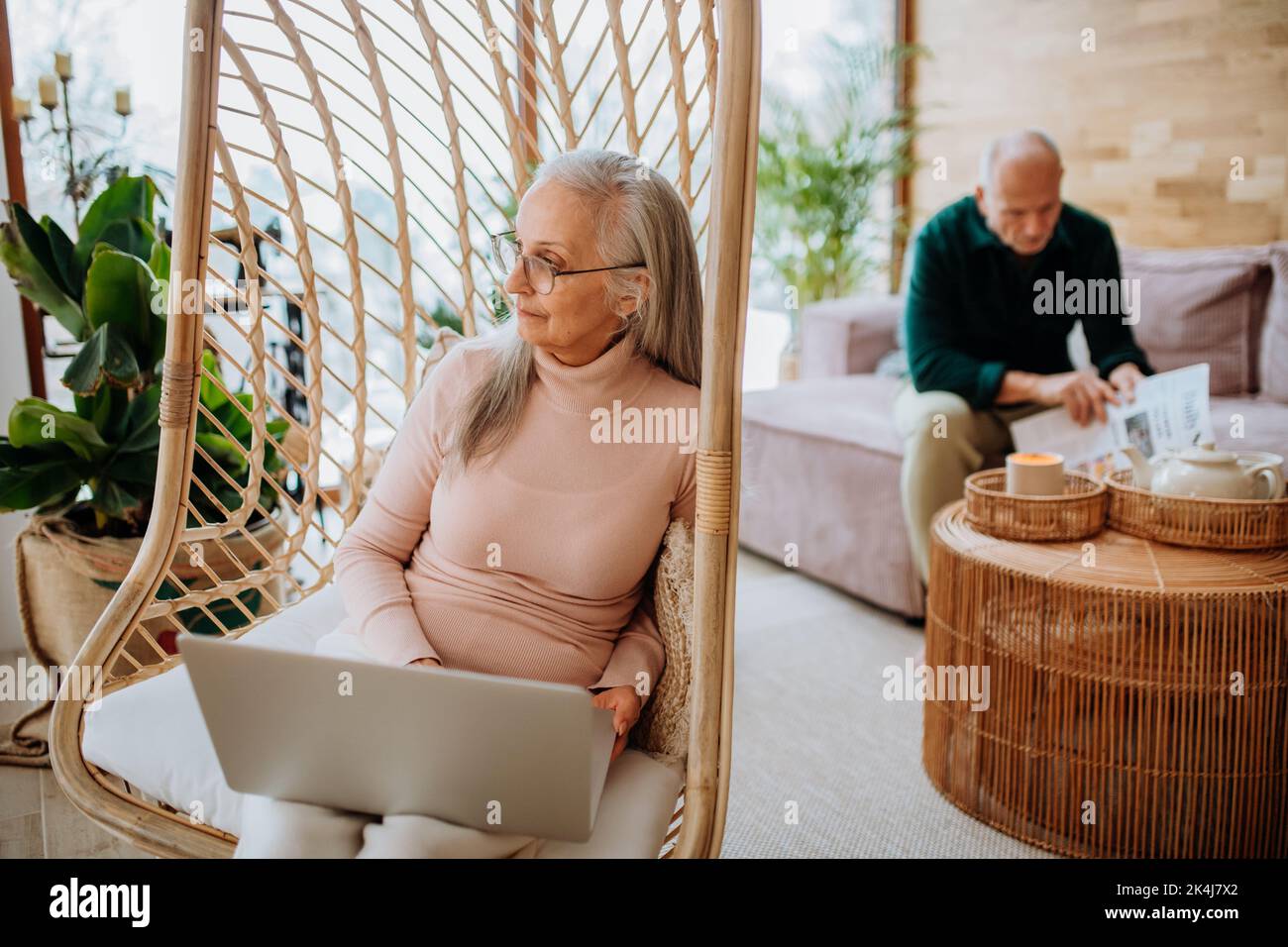 Senior woman sitting in outdoor swing and working at laptop, her husband resting at sofa. Concept of home office and active senior lifestyle. Stock Photo
