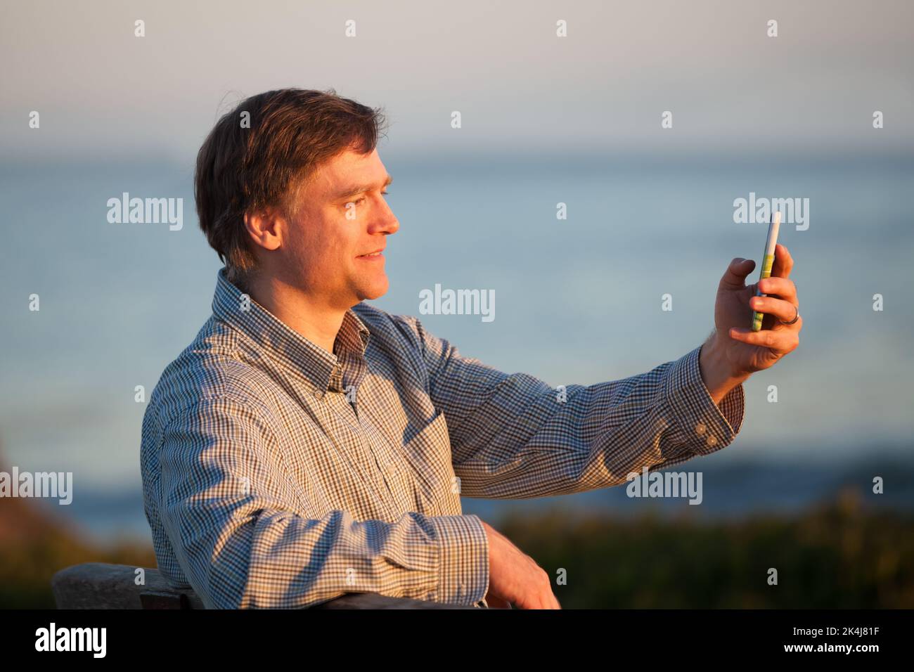 Caucasian man talking on smartphone while relaxing by ocean at sunset Stock Photo