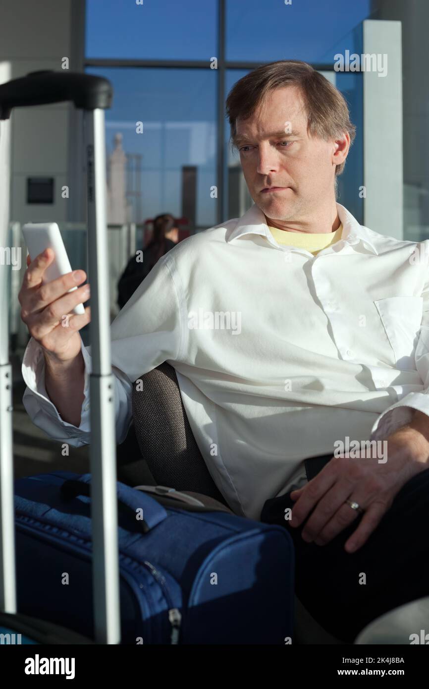 Caucasian businessman in fifties relaxing with legs up at airport terminal while using smartphone Stock Photo