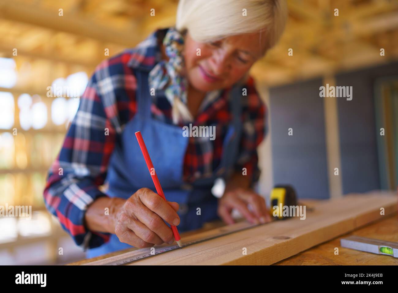 Senior woman measuring wooden board inside of her unfinished ecological wooden eco house. Stock Photo