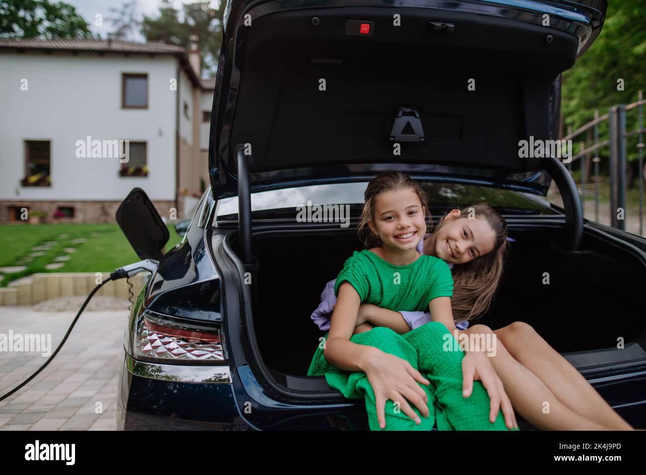 Happy sisters sitting in car trunk and waiting for electric car charging. Stock Photo