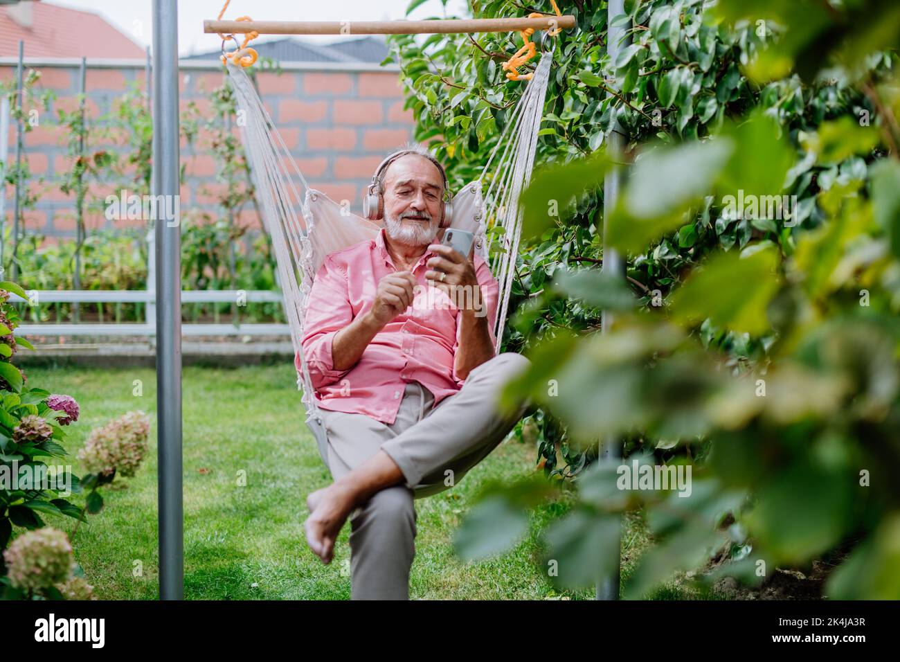 Happy senior man sitting in outdoor garden swing, listening music in headphones and scrolling smartphone. Stock Photo