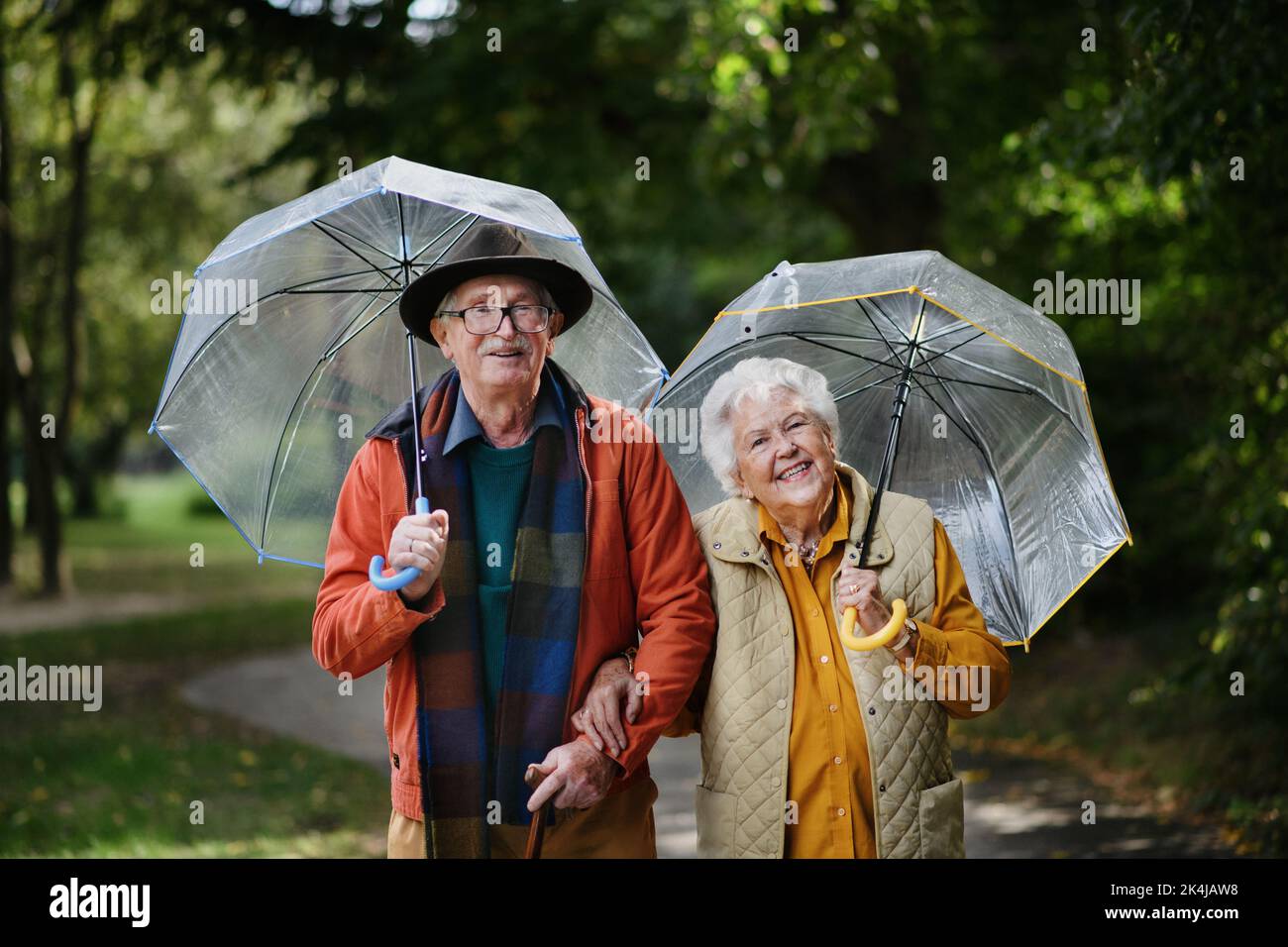 Happy senior couple walking with umbrellas in city park together. Stock Photo