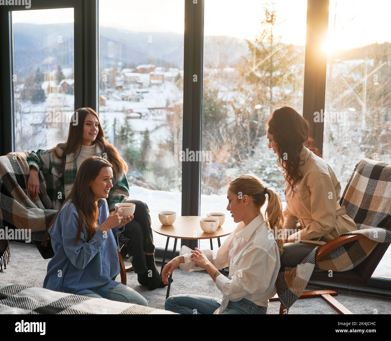 Young women enjoying winter weekends inside contemporary barn house. Four girls having fun and drinking hot tea near panoramic windows at sunset. Stock Photo