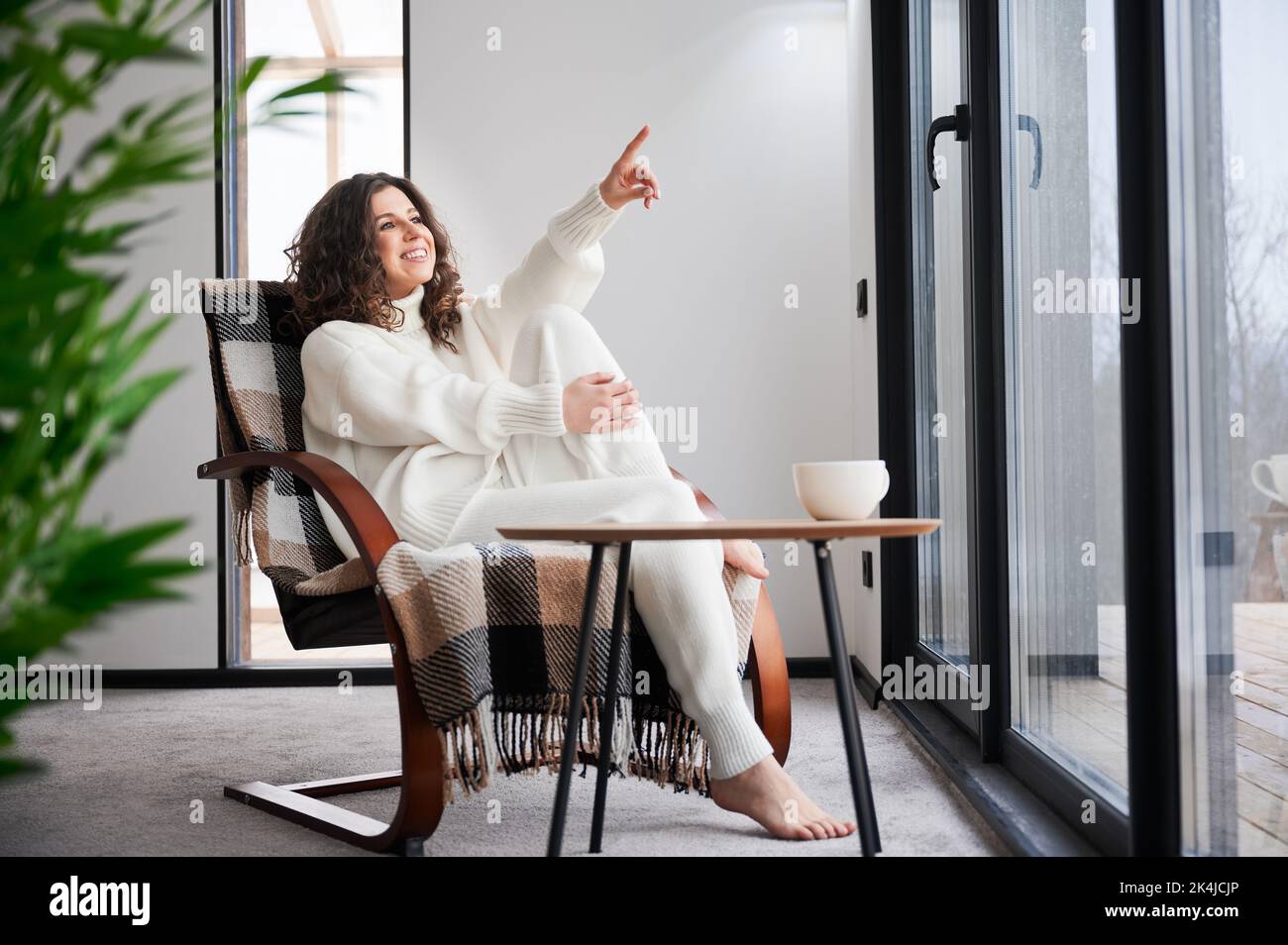 Curly young woman enjoying weekends inside contemporary barn house. Happy female tourist sitting on chair, pointing through panoramic windows in new cottage. Stock Photo