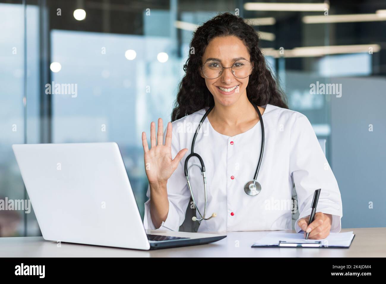 Portrait of a hispanic woman doctor veterinarian. He sits in the office, conducts an online consultation with a laptop, looks and waves at the camera. Records the diagnosis, treatment of the animal. Stock Photo