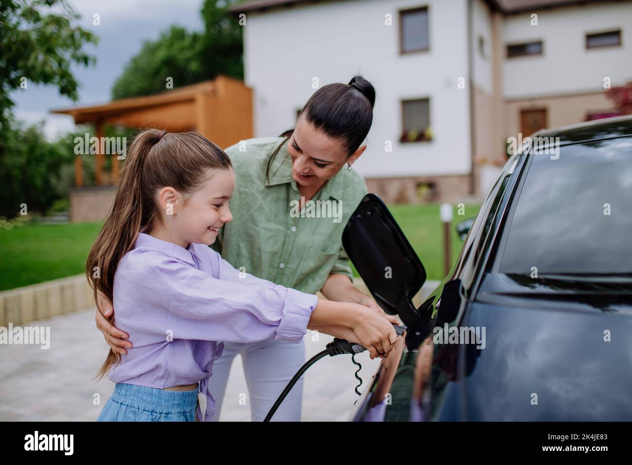 Happy mother showing her little daughter how to charge their electric car. Stock Photo
