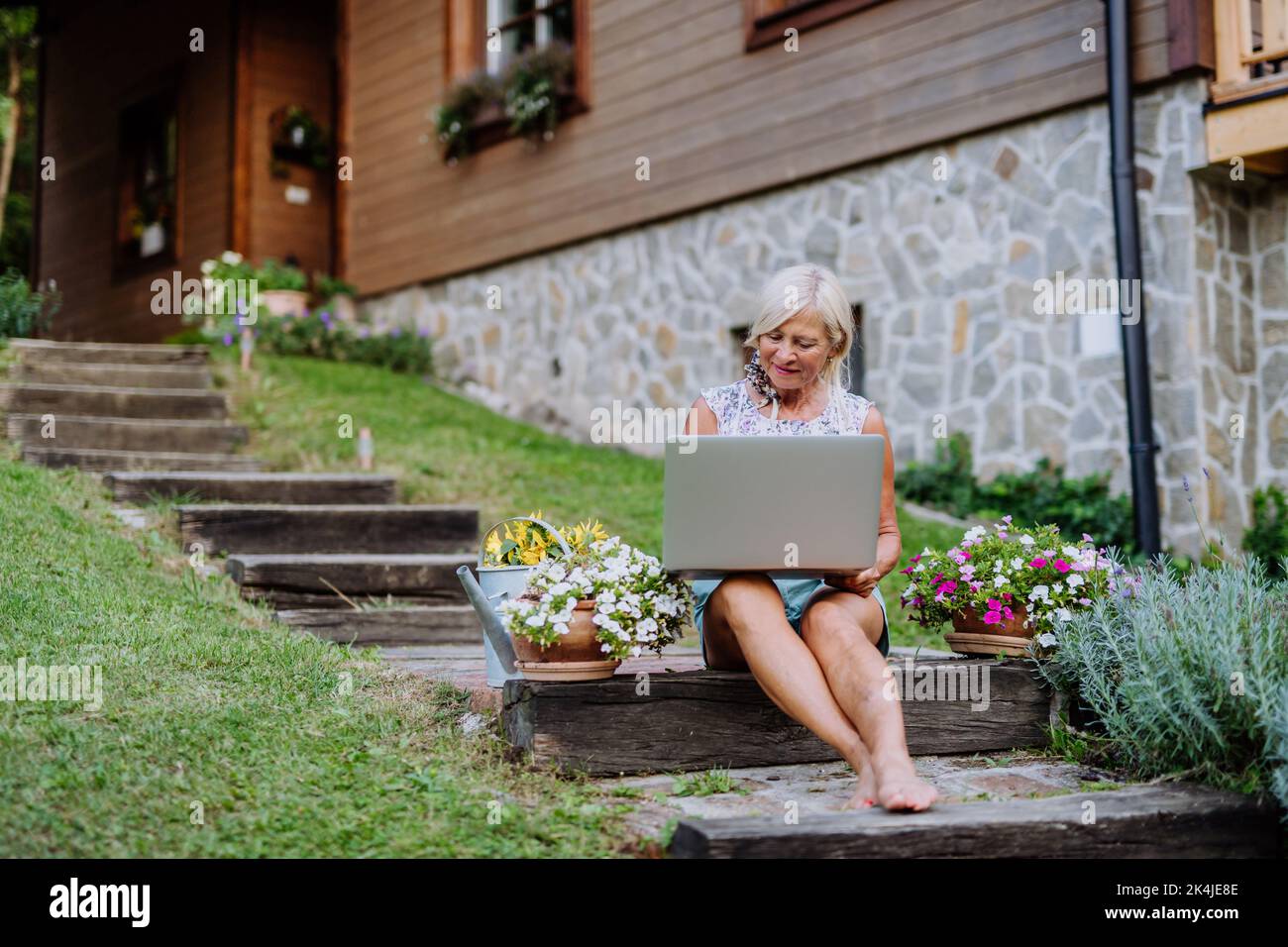 Senior woman using laptop and handling orders of her homegrown organic flowers and vegetables in garden, small business concept. Stock Photo