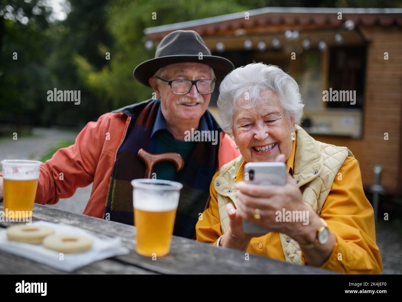 Happy senior couple in forest buffet resting after walk, having beer and looking at smartphone. Stock Photo