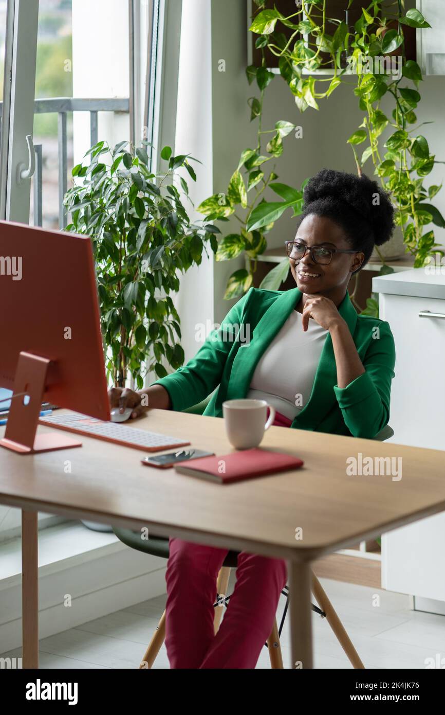 Smiling African American woman secretary working sits at computer desk office with green plants Stock Photo