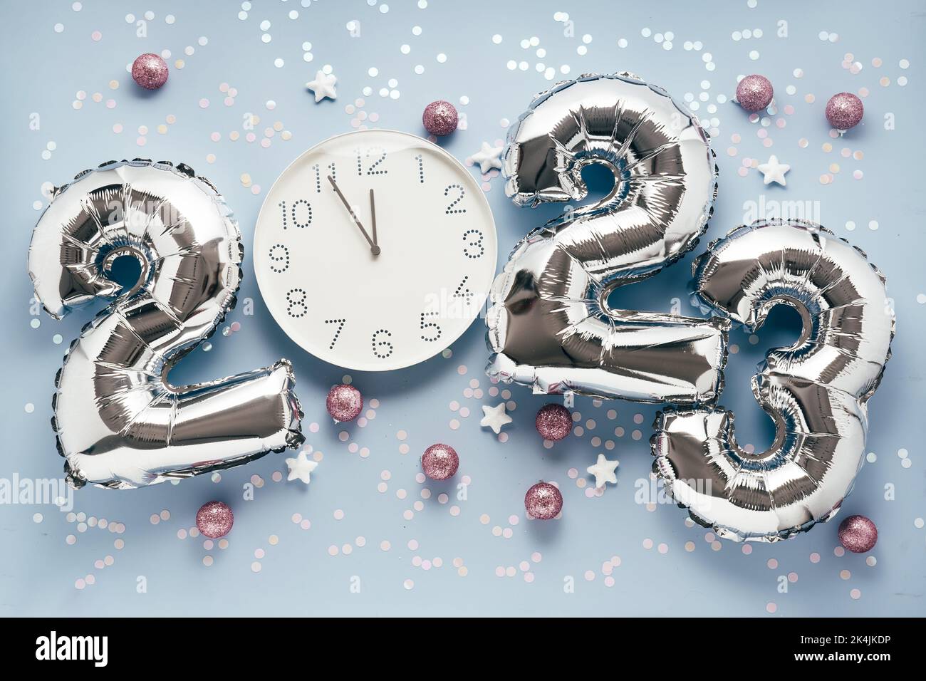 Shiny silber air balloon numbers and white watch at 12 o'clock on light blue background with confetti and decorations. 2023 New Year concept celebration Stock Photo