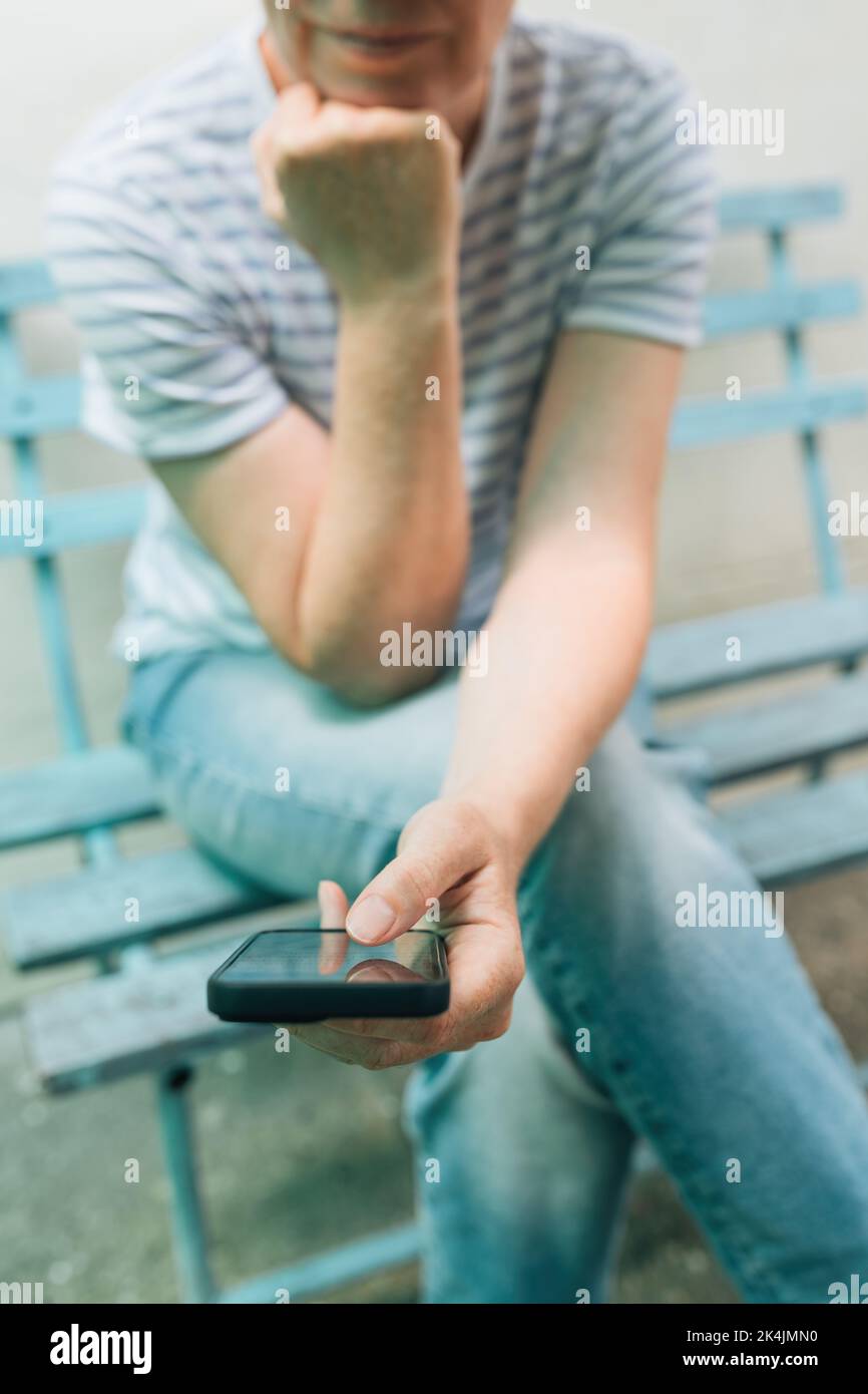 Woman typing text message on mobile smart phone while sitting on wooden bench. Female person wearing jeans trousers and stripped t-shirt using smartph Stock Photo