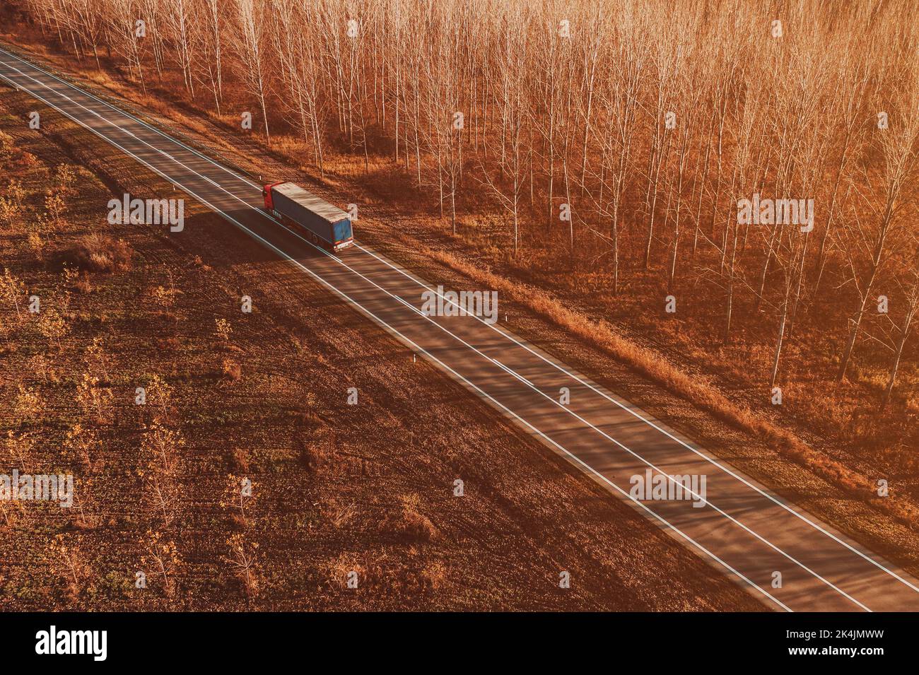 Semitrailer eighteen-wheeler truck on the road through deciduous forest landscape in autumn sunset, drone photography from above Stock Photo