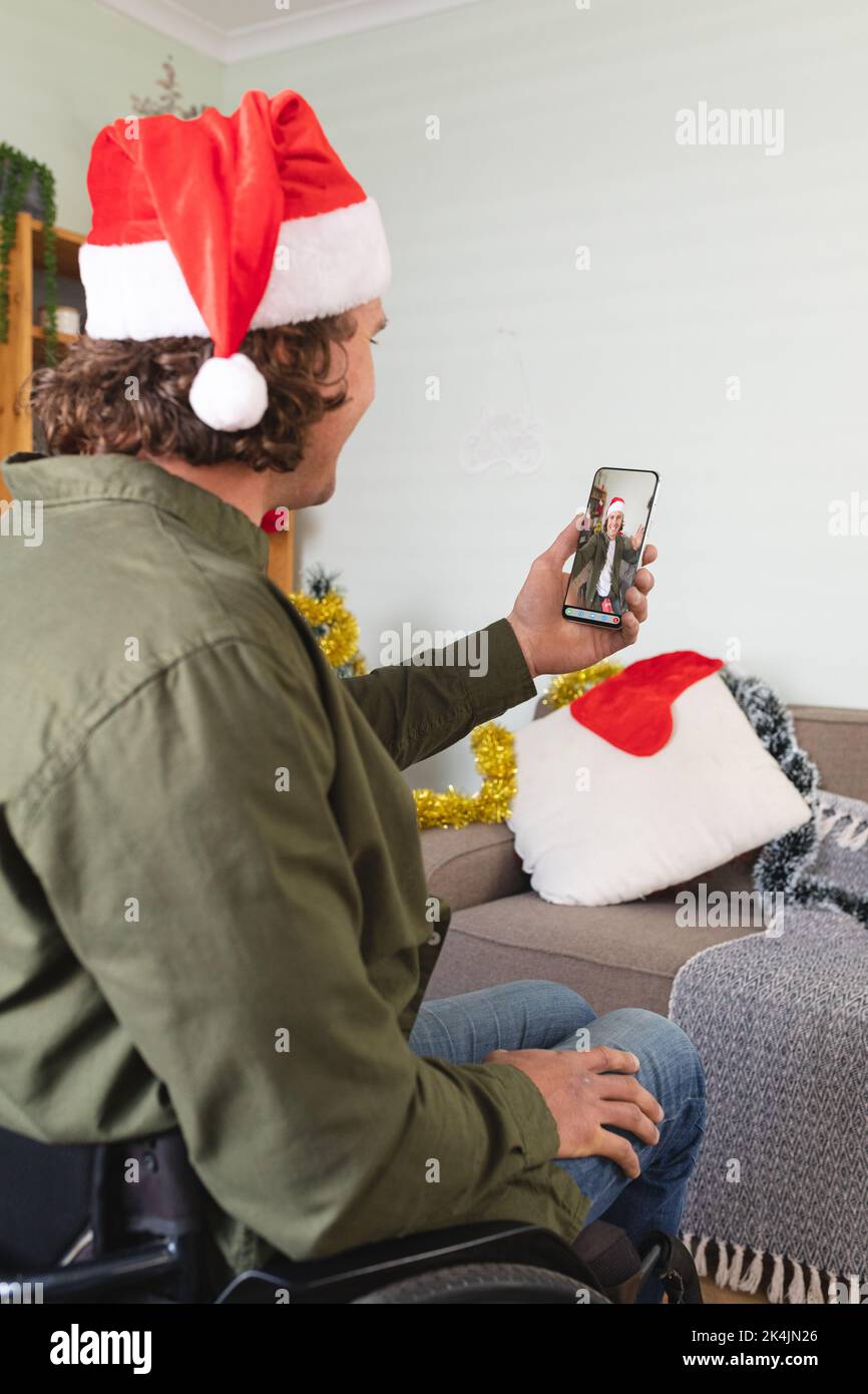 Caucasian man in wheelchair and santa hat making christmas smartphone video call with male friend. christmas, festivity and communication technology. Stock Photo