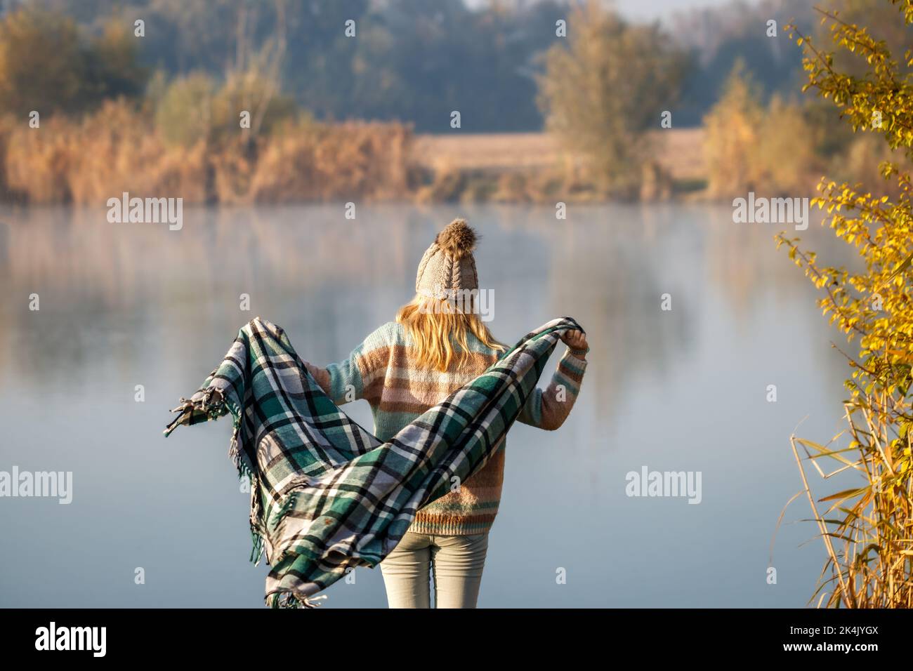 Woman with knit hat is wrapping herself in blanket at lake. Autumn camping in nature Stock Photo
