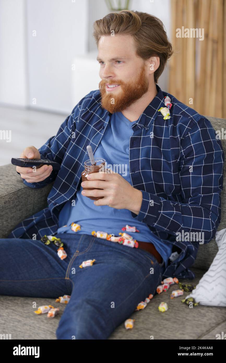 man covered in sweets on the sofa holding remote-control Stock Photo