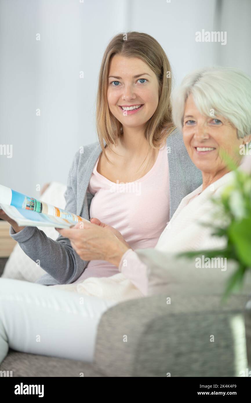 cheerful granddaughter and grandmother looking through magazine sat on sofa Stock Photo
