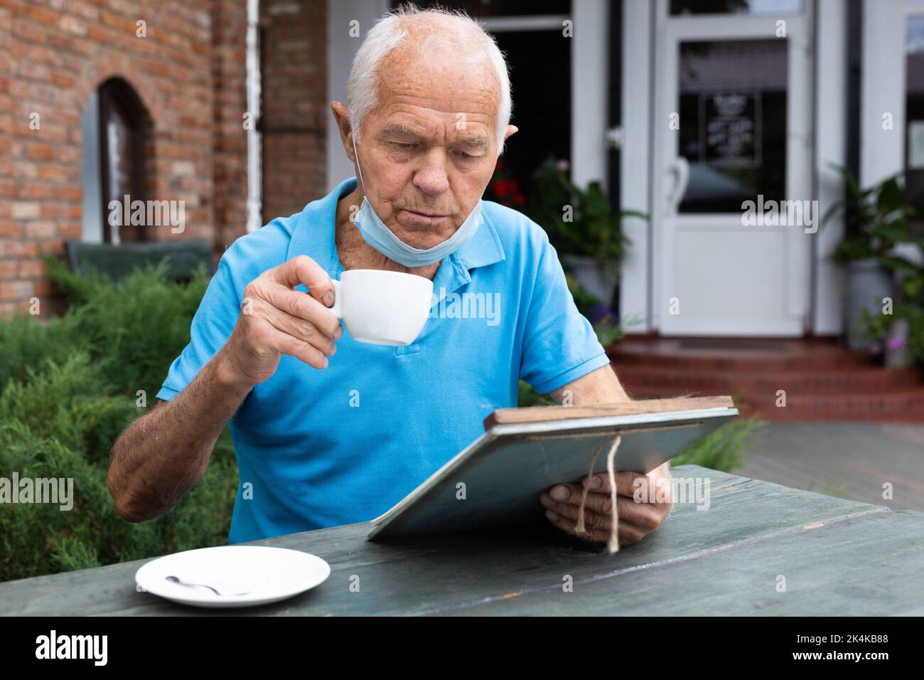 Senior man in protective mask chooses dish from the menu while sitting at the table of street cafe Stock Photo