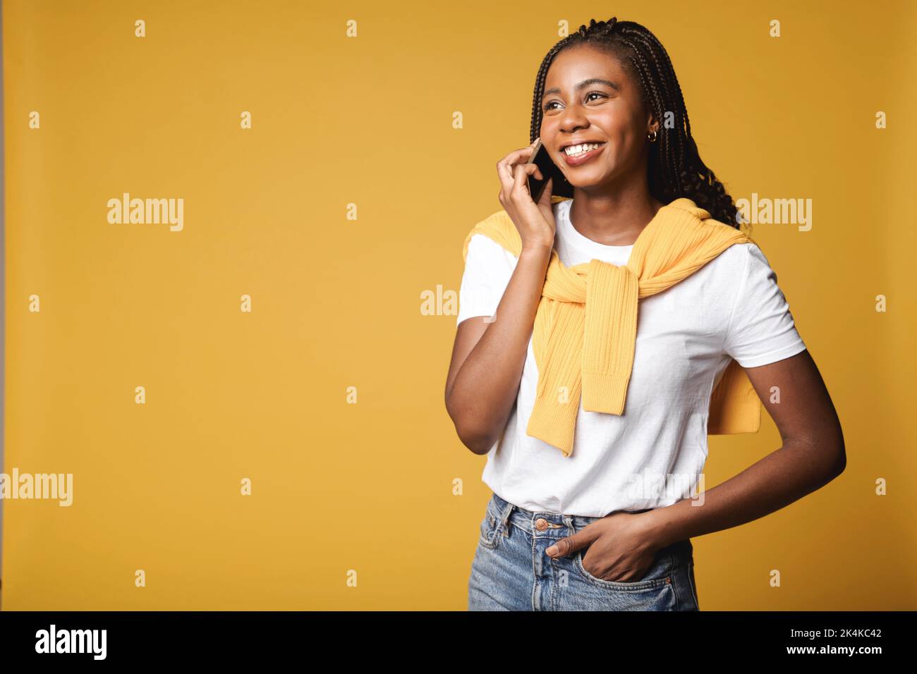 Cheerful smiling woman talking on smartphone and looking away while standing isolated on yellow background. Multiracial lady has pleasant phone conversation Stock Photo